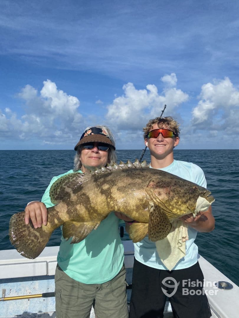A young angler and an adult proudly display a large Goliath Grouper while fishing in clear waters, showcasing a successful day on the boat.