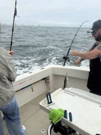 Two anglers actively fishing on a boat, with one reeling in a catch while the other prepares their line, showcasing a dynamic fishing experience.