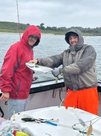 Two anglers proudly displaying a large Striped Bass while fishing on a boat in overcast weather.