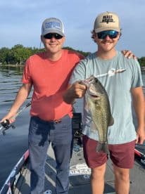 Two anglers proudly displaying a large Largemouth Bass while fishing on a serene lake, showcasing a successful day on the water.