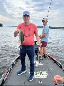 An adult angler proudly displaying a Largemouth Bass while fishing on a boat, showcasing a scenic lake environment.