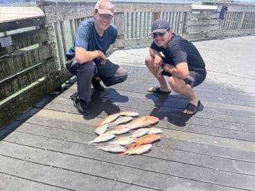 Two anglers posing with a variety of fish, including Snapper, laid out on the dock after a successful fishing trip.