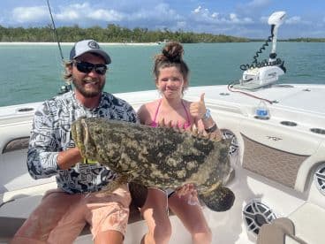 A young angler and an adult proudly displaying a large Goliath Grouper on a boat in a scenic fishing environment.
