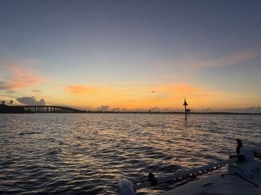 A picturesque sunset view from the side of a boat, showcasing the tranquil waters and a bridge in the background, perfect for evening fishing trips.