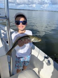 Young angler proudly holding a Speckled Trout while fishing from a boat in calm waters.