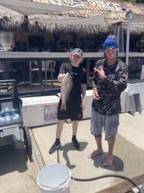 A young angler proudly displaying a large Snapper alongside an experienced fisherman at a dockside restaurant after a successful fishing trip.