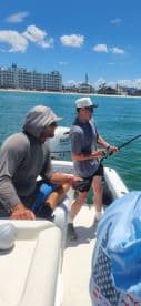 Two anglers actively fishing from a small boat, with a sunny coastal backdrop and clear blue waters.