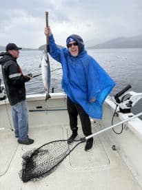 Enthusiastic angler holding a large Salmon while fishing on a boat in a scenic coastal environment.
