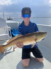 Young angler proudly holding a large Redfish while fishing on a boat in a coastal environment.