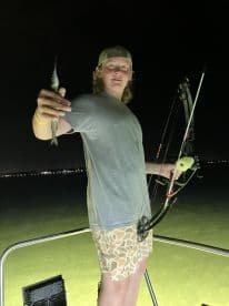 A young angler proudly displaying a small fish caught at night while bowfishing from a boat, with city lights in the background.
