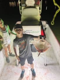 A young angler proudly displaying a caught stingray on a fishing boat at night, with a family member in the background.