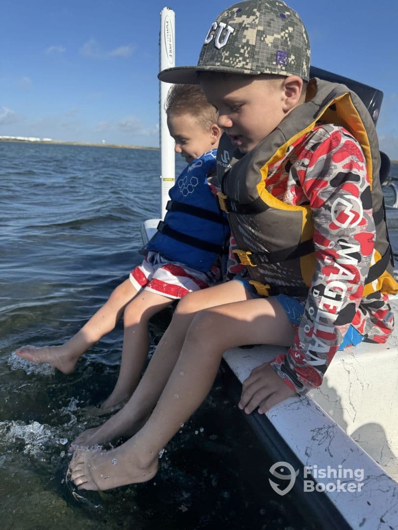 Two young boys enjoying a day on the water, sitting on the edge of a boat with their feet in the water, showcasing a fun fishing experience.