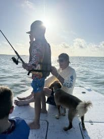 A young boy in a life jacket is casting a fishing line while enjoying a sunny day on the water, accompanied by an adult and a dog.