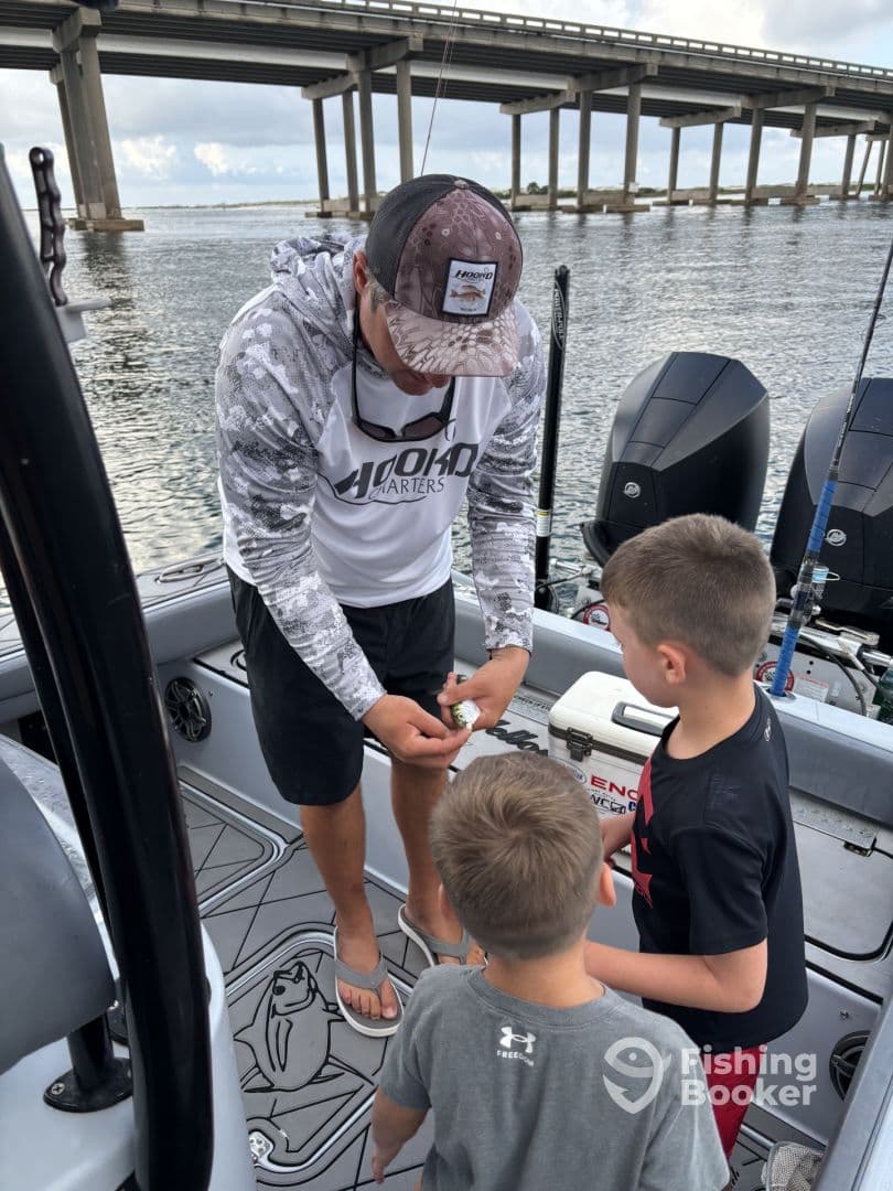 A fishing guide demonstrates bait preparation to two young anglers on a boat, with a bridge in the background, highlighting an engaging fishing experience.
