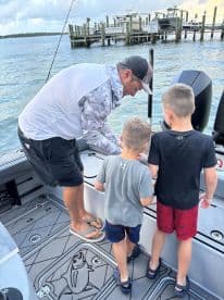 A fishing guide interacts with two young anglers on the deck of a boat, preparing for a day of fishing in a scenic coastal environment.