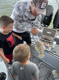 A young angler excitedly observes a Flounder being held by an adult on a fishing boat, highlighting a fun family fishing experience.