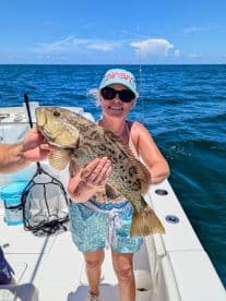 A woman proudly displaying a large Grouper while fishing on a boat in sunny waters.