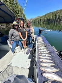 A group of anglers posing on a boat with a neatly arranged catch of trout displayed on a towel, showcasing a successful fishing trip in a scenic lake environment.