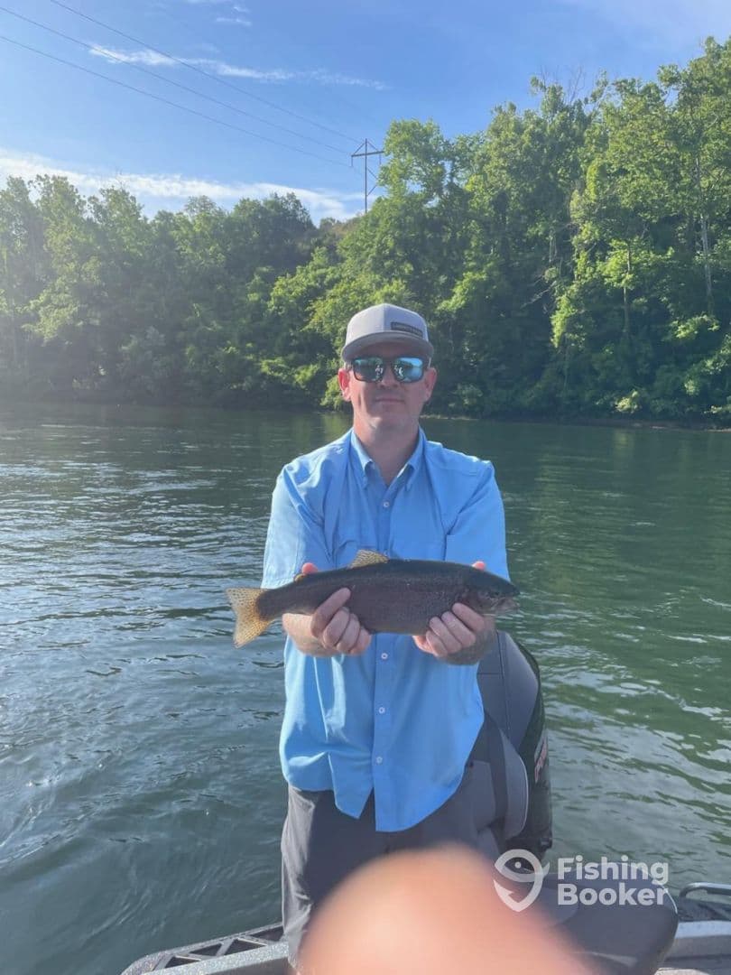 Angler proudly displaying a Rainbow Trout while fishing on a serene river, surrounded by lush greenery.