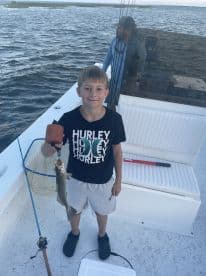 A young angler proudly displaying a Speckled Trout while fishing on a boat, showcasing a fun family outing.