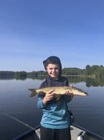 A young angler proudly displaying a Northern Pike while fishing on a calm lake, showcasing a beautiful day outdoors.
