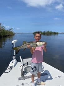 A young angler proudly displaying a Snook while fishing from a boat in a calm waterway, showcasing a successful day on the water.