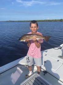 A young angler proudly displaying a Redfish while fishing from a boat in a scenic waterway.