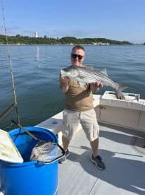 An angler proudly displaying a striped bass while fishing on a boat, surrounded by a scenic waterfront.