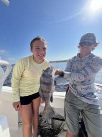 Two young anglers proudly displaying a Black Drum caught during a fishing trip on a boat.