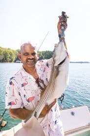 An angler proudly displaying a large striped bass while fishing on a serene lake, showcasing a vibrant summer outfit.