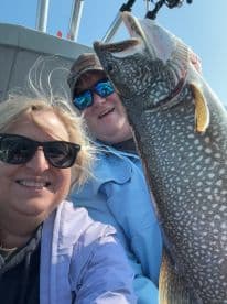 Two anglers smiling for a selfie while proudly displaying a large Lake Trout caught during a fishing trip.