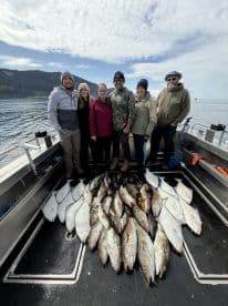 A group of anglers proudly posing with a large catch of Halibut and Cod on a fishing charter in a scenic coastal environment.