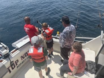 A young angler in a life jacket is reeling in a fish while accompanied by family members on a fishing charter in open waters.