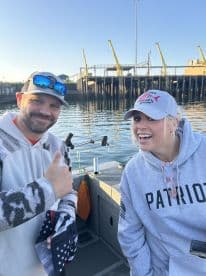 Two anglers smiling on a boat, enjoying a fishing trip with a scenic waterfront backdrop.