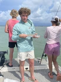 A young angler proudly displaying a speckled trout while fishing on a boat, with friends engaged in the activity in the background.
