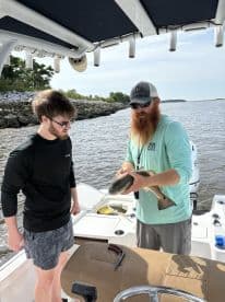 Two anglers on a boat examining a caught fish, likely a Redfish, in a scenic waterway.