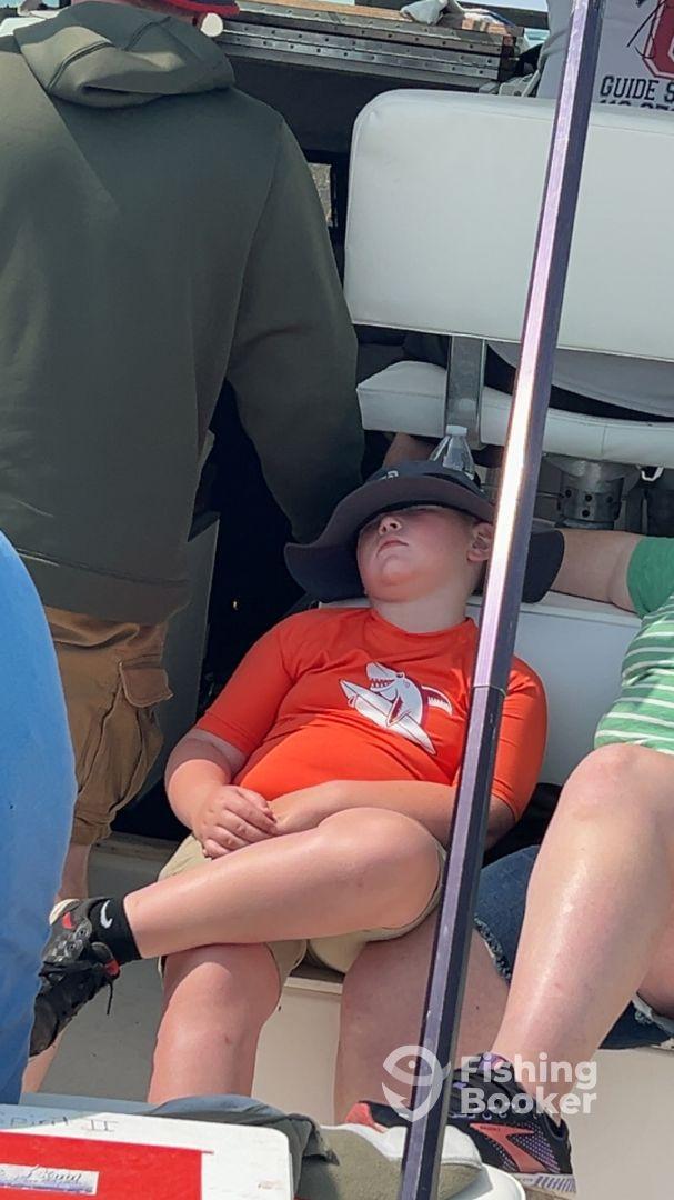 A young boy resting in a seated position on a fishing boat, wearing a shark-themed shirt and a wide-brimmed hat.