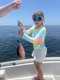 A young angler proudly displaying a Snapper while fishing on a boat in clear blue waters.