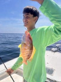 A young angler proudly displaying a colorful Snapper while fishing on a sunny day at sea.