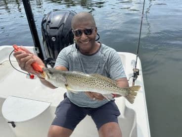 Angler proudly displaying a large Speckled Trout while fishing from a small boat in calm waters.