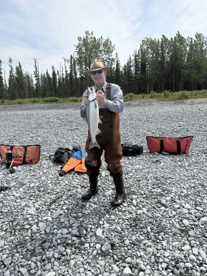 An angler proudly displaying a large fish on a rocky riverbank, surrounded by fishing gear and lush greenery in the background.