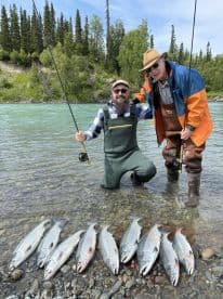 Two anglers proudly displaying their catch of silver salmon along a scenic riverbank, showcasing a successful day of fishing in a beautiful natural setting.