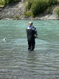 Angler wading in a river, casting a line while fishing in a scenic environment surrounded by lush greenery.