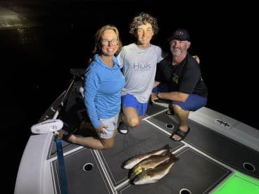 A family poses on a boat at night, proudly displaying their catch of three Redfish after a successful fishing trip.