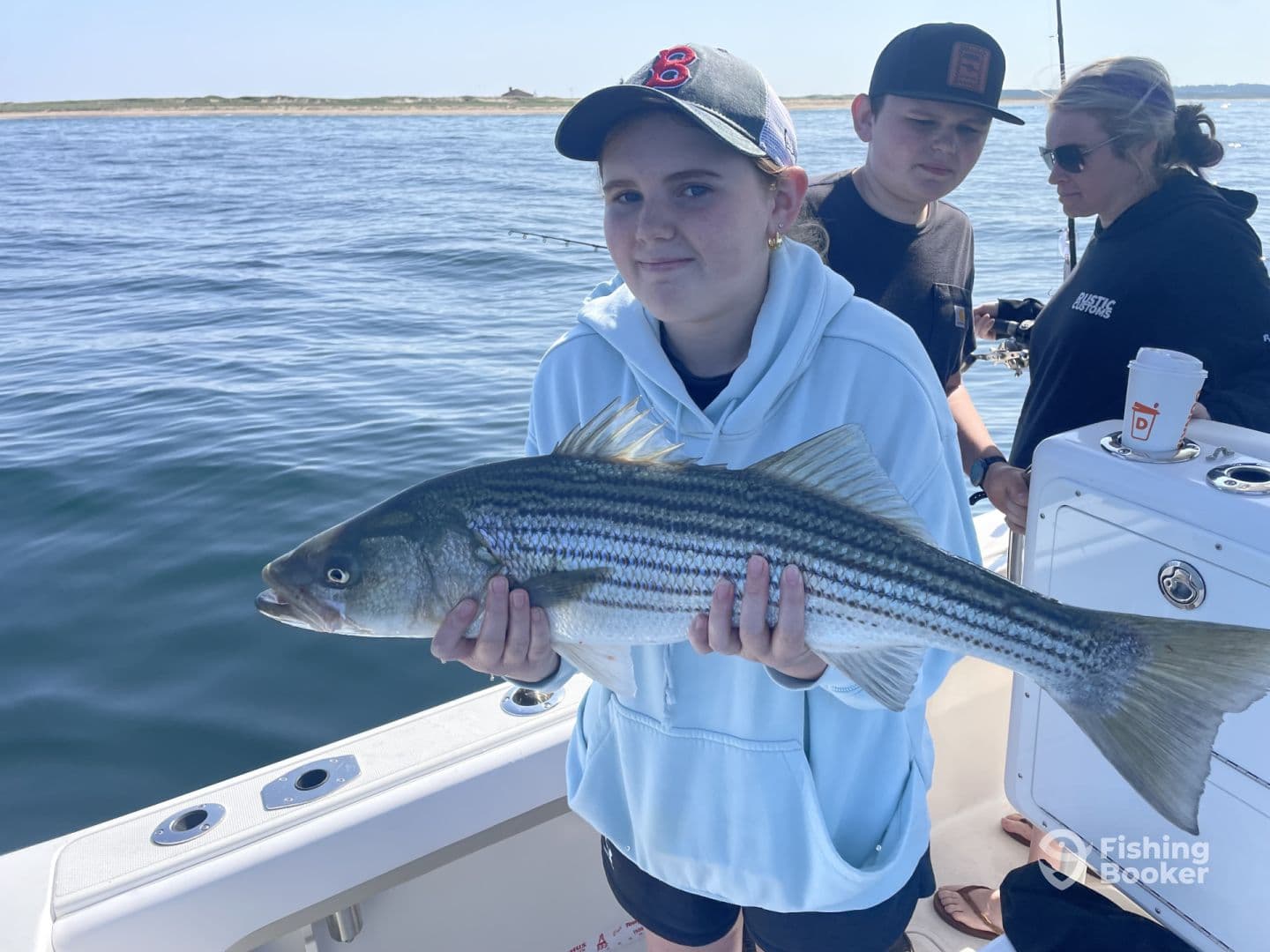 A young angler proudly displaying a large Striped Bass while fishing on a boat, surrounded by fellow anglers.