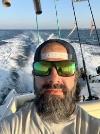 Selfie of an angler on a boat, showcasing fishing rods in the background while cruising on open water.