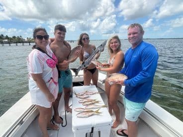 A group of four anglers proudly displaying their catch, including a large Barracuda and Snapper, aboard a boat in a sunny coastal setting.