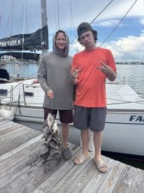 Two young anglers proudly displaying their catch of fish at the dock, with a sailboat in the background.