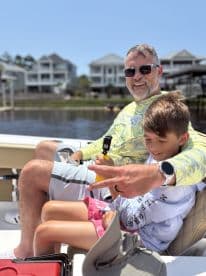 A father and son enjoying a sunny day on a boat, showcasing a relaxed atmosphere with waterfront homes in the background.