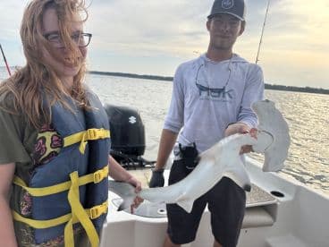 A young angler wearing a life jacket proudly displays a caught Hammerhead Shark while on a boat, showcasing a successful fishing adventure.